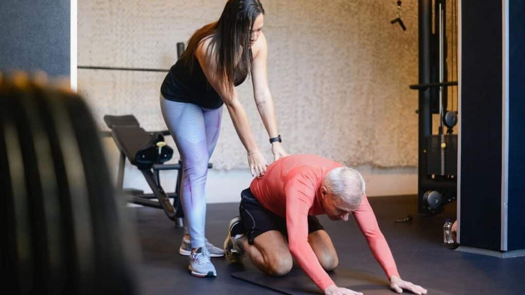 Elderly man assisted by trainer during a workout session inside a gym setting, emphasizing healthy living.