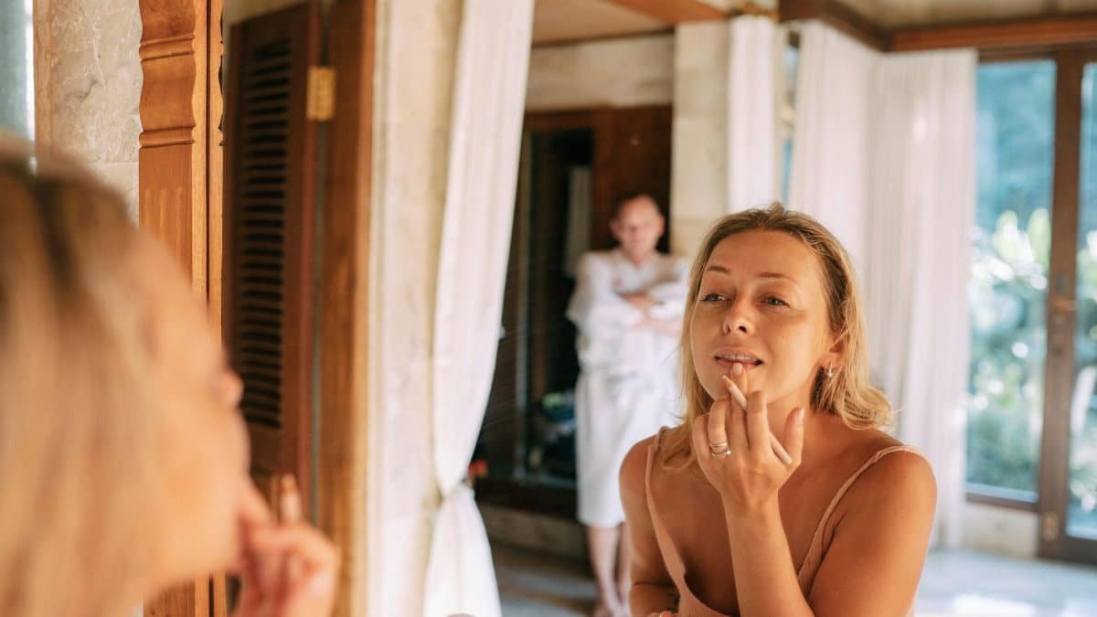 A woman applies makeup in a luxurious bathroom as a man relaxes in the background.