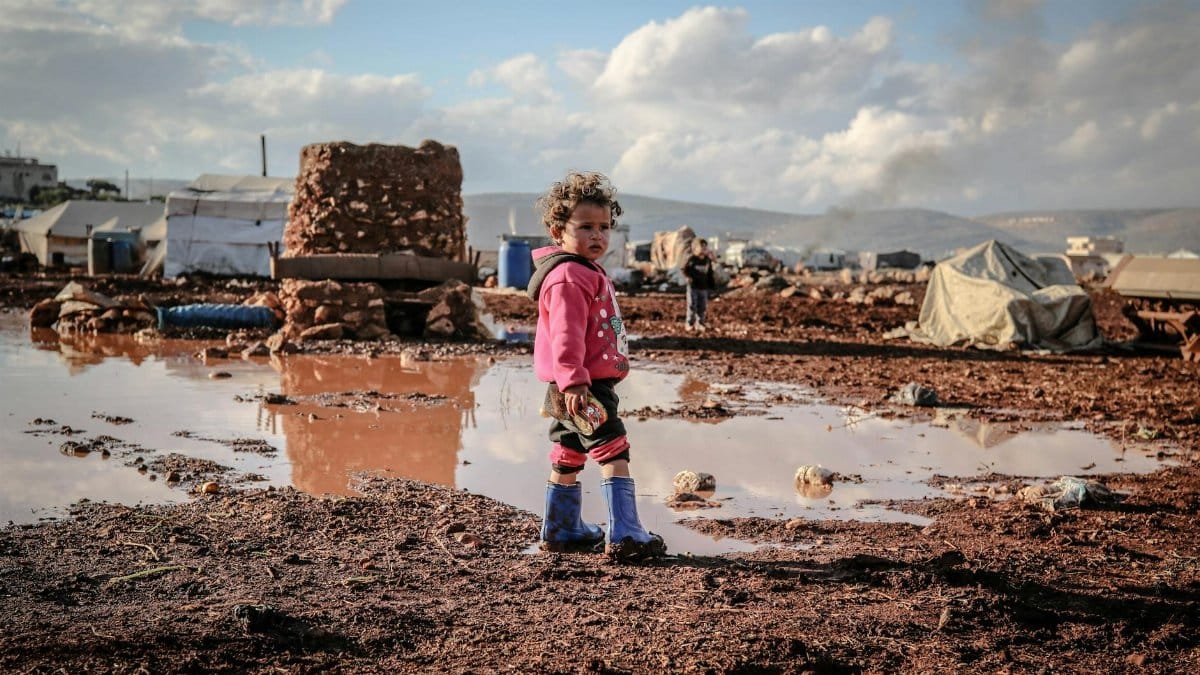 A young child stands in a muddy refugee camp in Idlib, Syria, reflecting resilience amidst adversity.