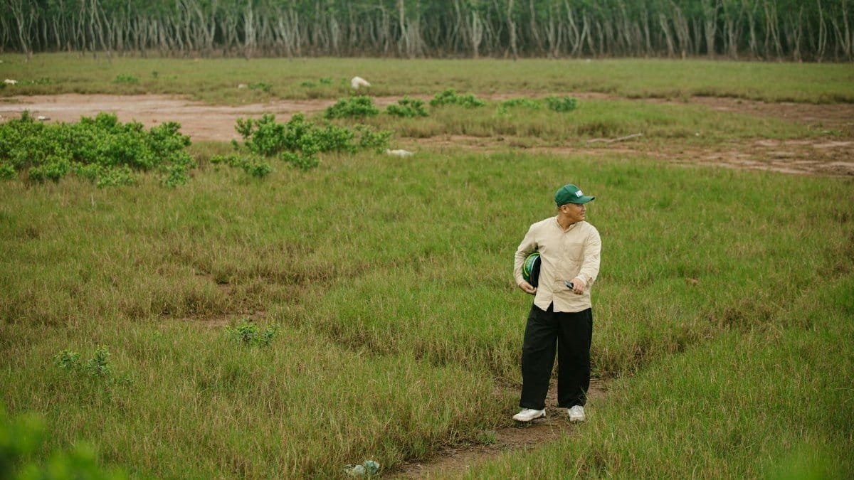 A man in casual attire walking through a lush green field with trees in the background.