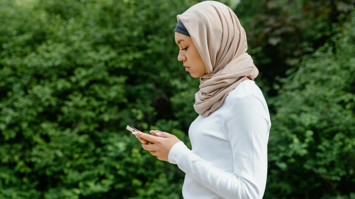 Side view of a Muslim woman in hijab using a smartphone in a natural setting.