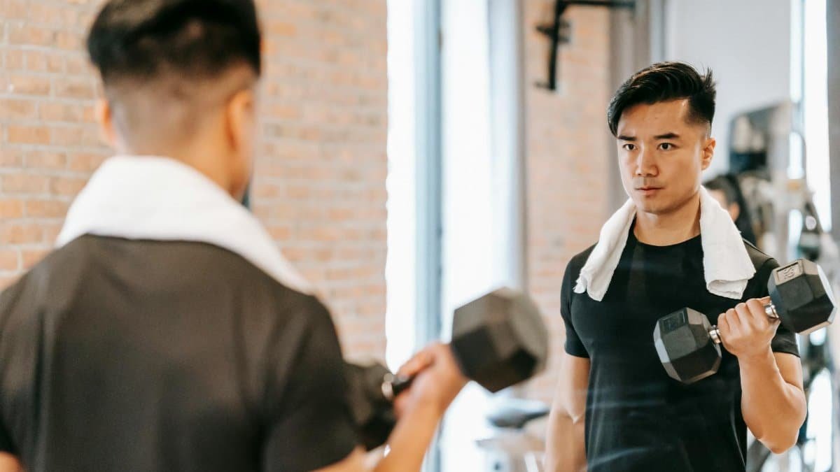 Active Asian male with towel lifting heavy dumbbell and looking in mirror while exercising in modern gym with brick wall