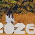 A person sitting outdoors with large numbers spelling 2026, surrounded by tropical foliage.