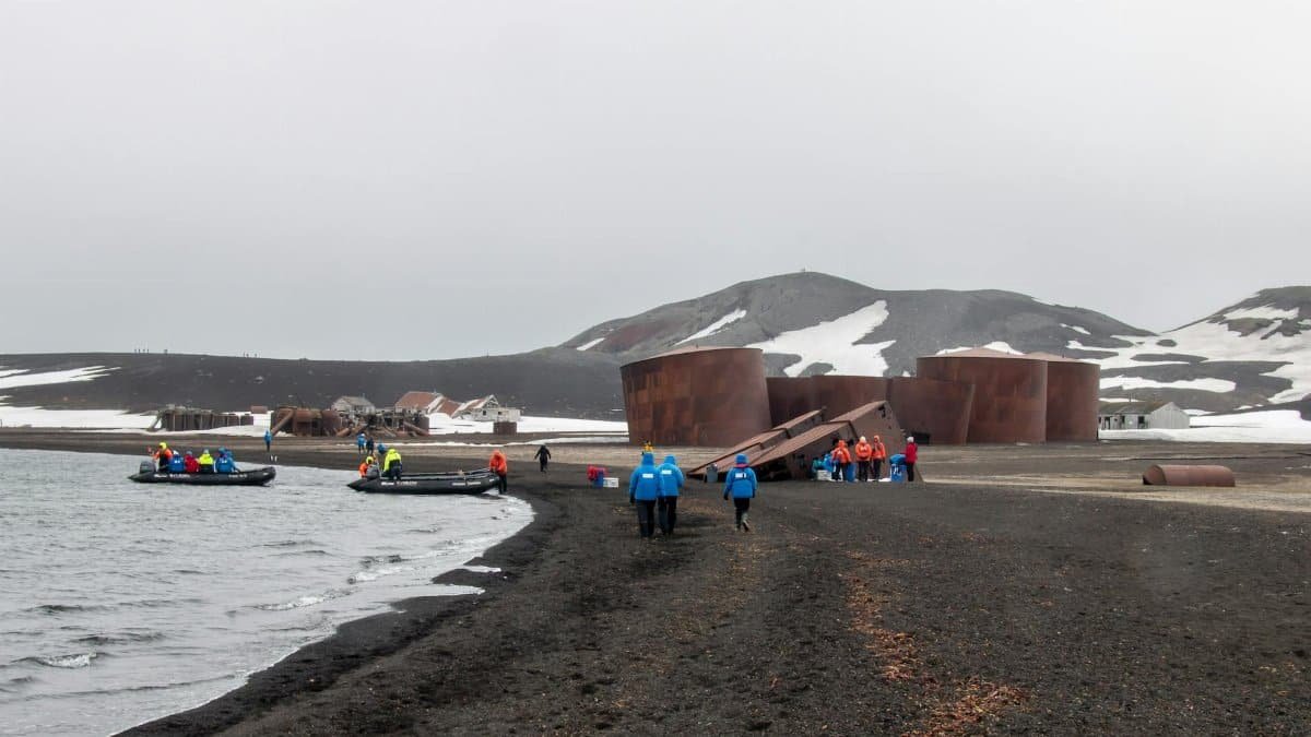 Explorers arrive by boat at Deception Island, Antarctica, with historical ruins and snowy landscapes.