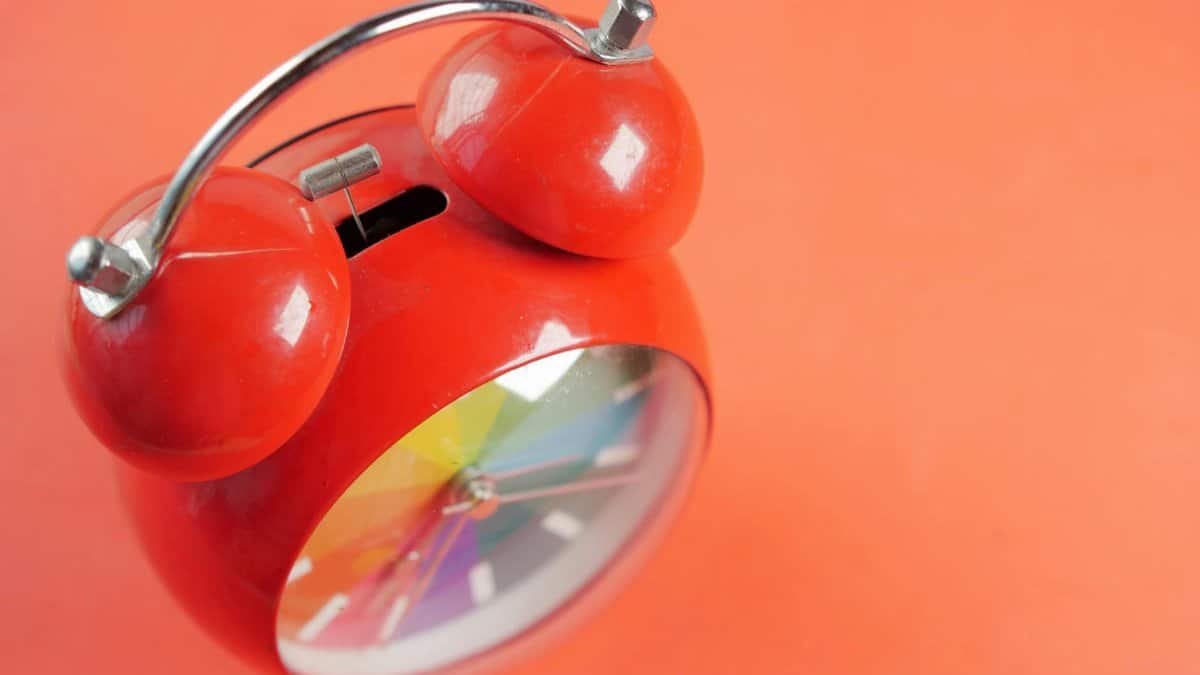 Close-up of a red analog alarm clock with a rainbow clock face on a bright orange background.