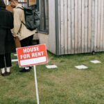 Group of adults viewing a wooden house with a 'House for Rent' sign on the lawn.