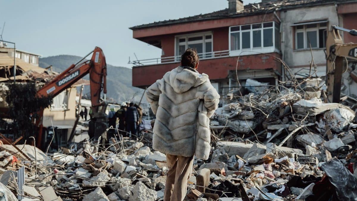 A woman in a fur coat stands amidst earthquake rubble, observing damaged buildings and an excavator.