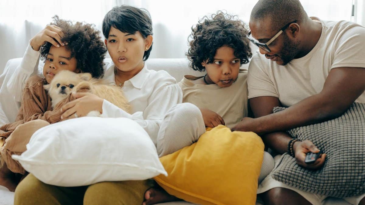Multiracial family resting on comfortable sofa together with little dog while mother and little son watching TV with interest and father and elderly son talking to each other