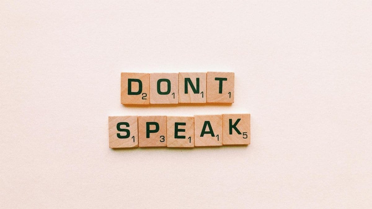 Wooden Scrabble tiles spelling 'Don't Speak' on a pink background, emphasizing silence.