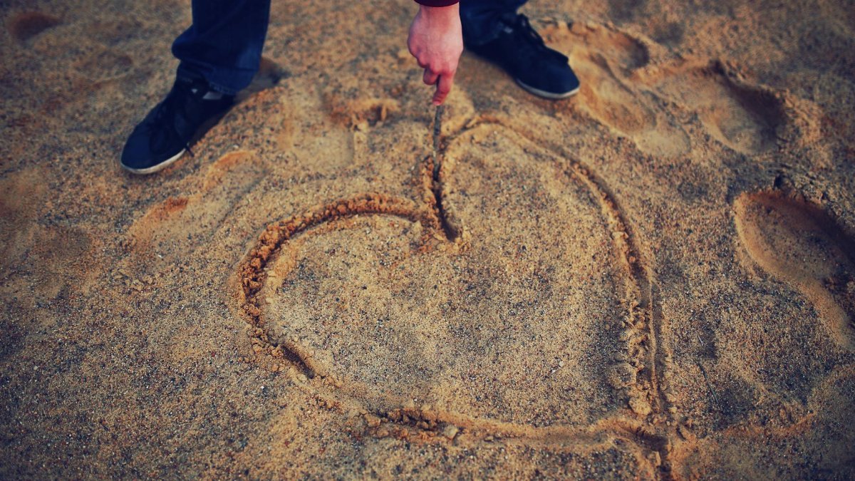 Person drawing a heart shape in sand, evoking romance and creativity.