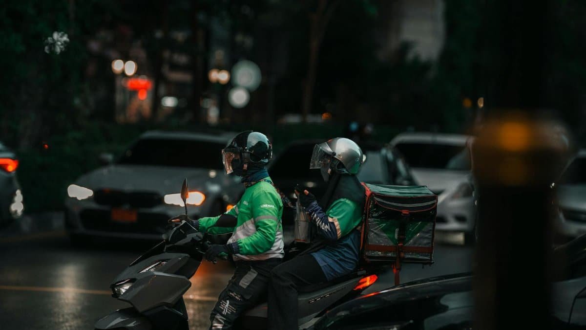 Two delivery riders on a motorbike navigating through city streets at night.
