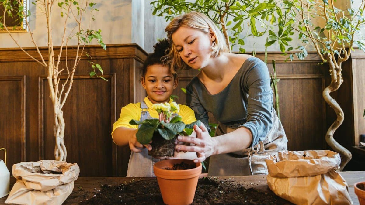 A woman and child enjoy planting a flower together indoors, surrounded by green plants.
