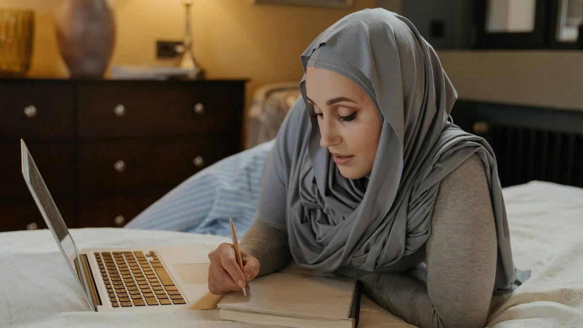 A Muslim woman in a hijab studying on her bed with a laptop and taking notes. Ideal for education and work-from-home visuals.