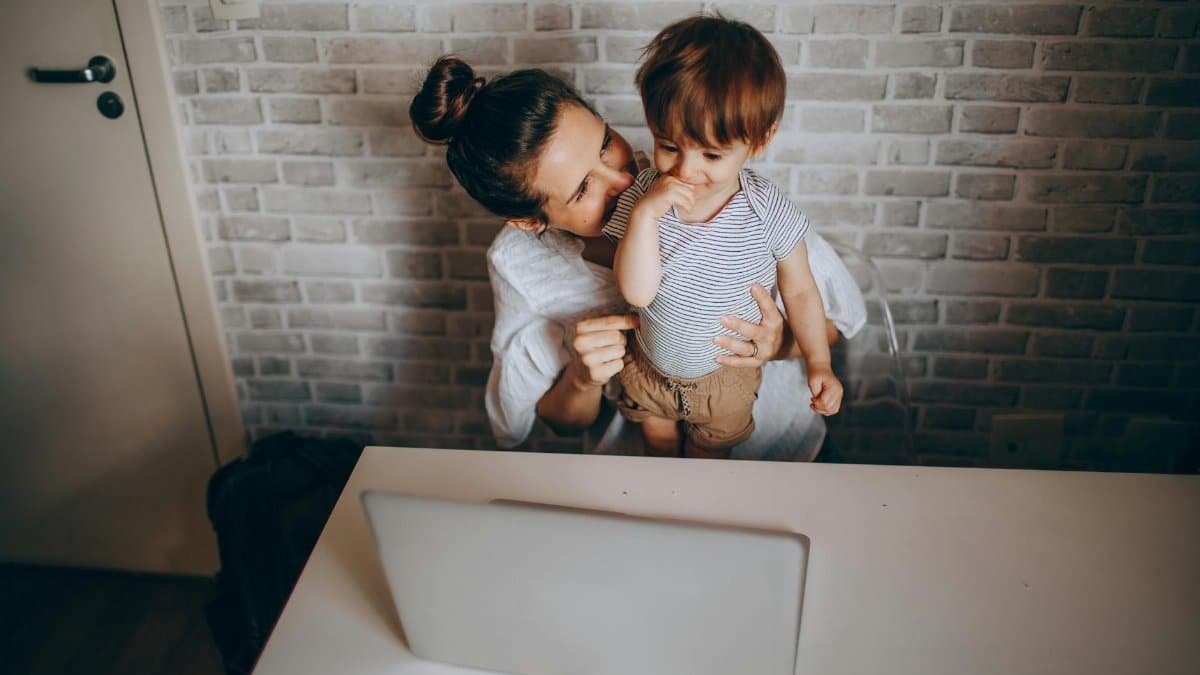 A mother lovingly interacts with her toddler while working on a laptop in a cozy home setting.