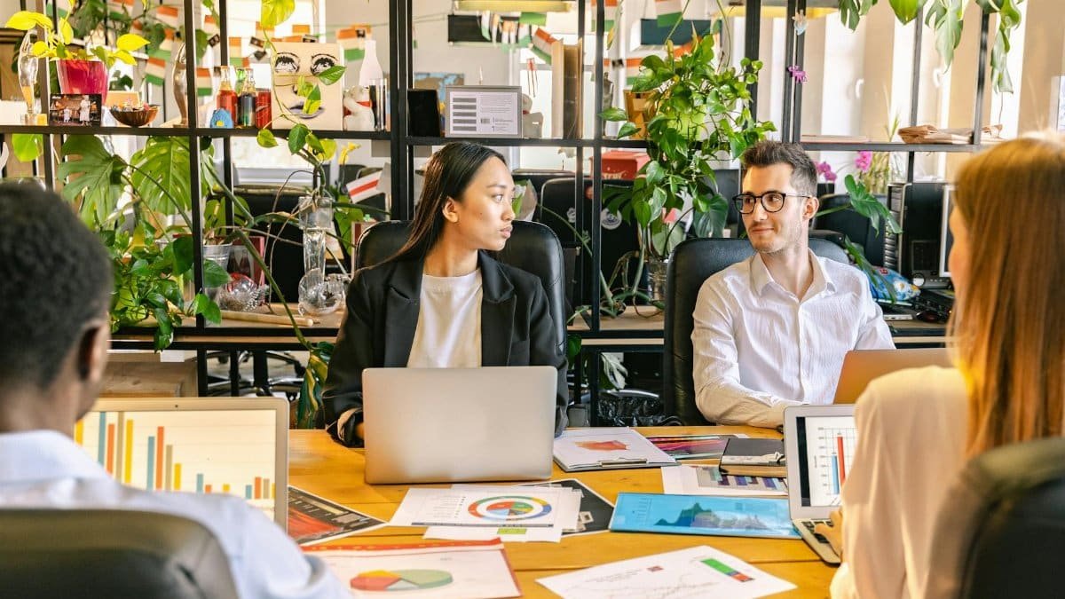 A diverse team of professionals discussing projects in a plant-filled office setting.