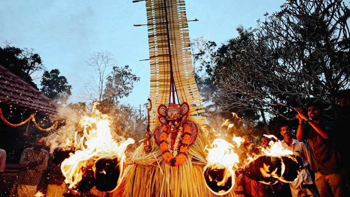 A colorful Theyyam dancer performs a fire ritual outdoors at twilight.