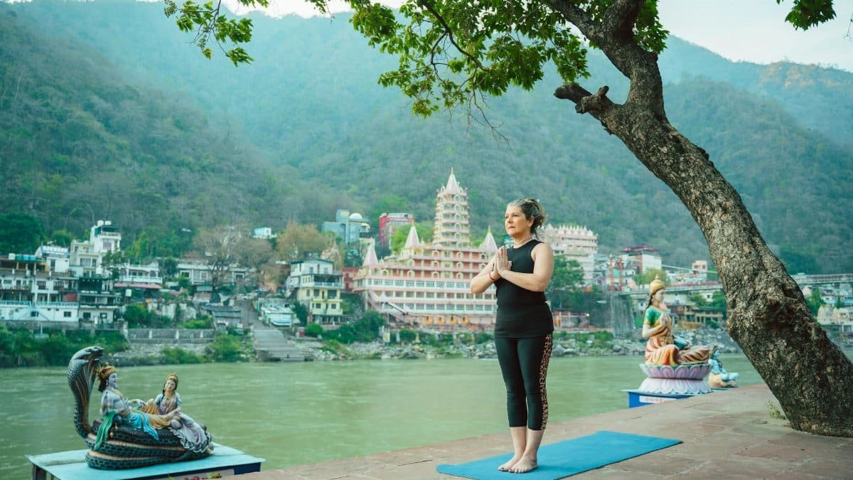 Woman practicing yoga by a serene riverside with statues in the backdrop, Rishikesh, India.