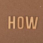Wooden letters spelling 'HOW' on brown textured cardboard background.