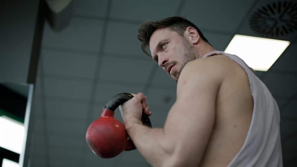 Low angle of adult male athlete wearing sportswear doing swing exercise with kettlebell in hand while training in sport center and looking over shoulder