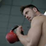 Low angle of adult male athlete wearing sportswear doing swing exercise with kettlebell in hand while training in sport center and looking over shoulder