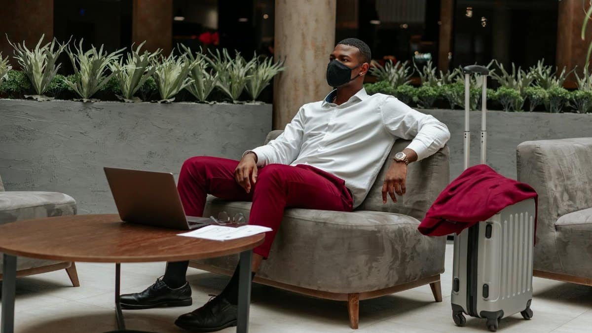 Man wearing facemask, sitting in hotel lobby with laptop and suitcase, adapting to new normal.