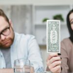 An adult interracial couple expressing concern about finances as they hold a one dollar bill indoors.