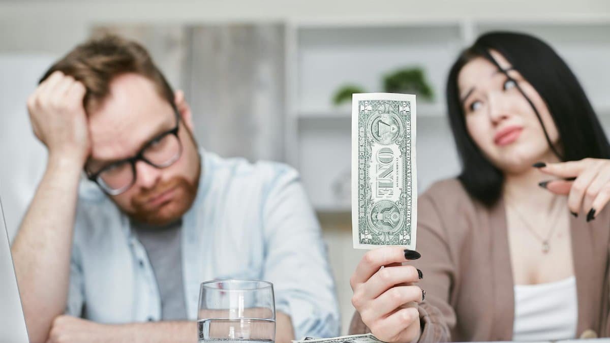 An adult interracial couple expressing concern about finances as they hold a one dollar bill indoors.