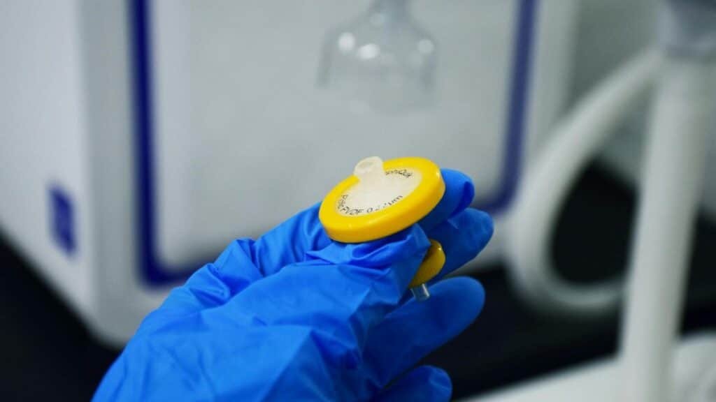 Detailed close-up of a hand in a blue glove holding a laboratory syringe filter indoors.