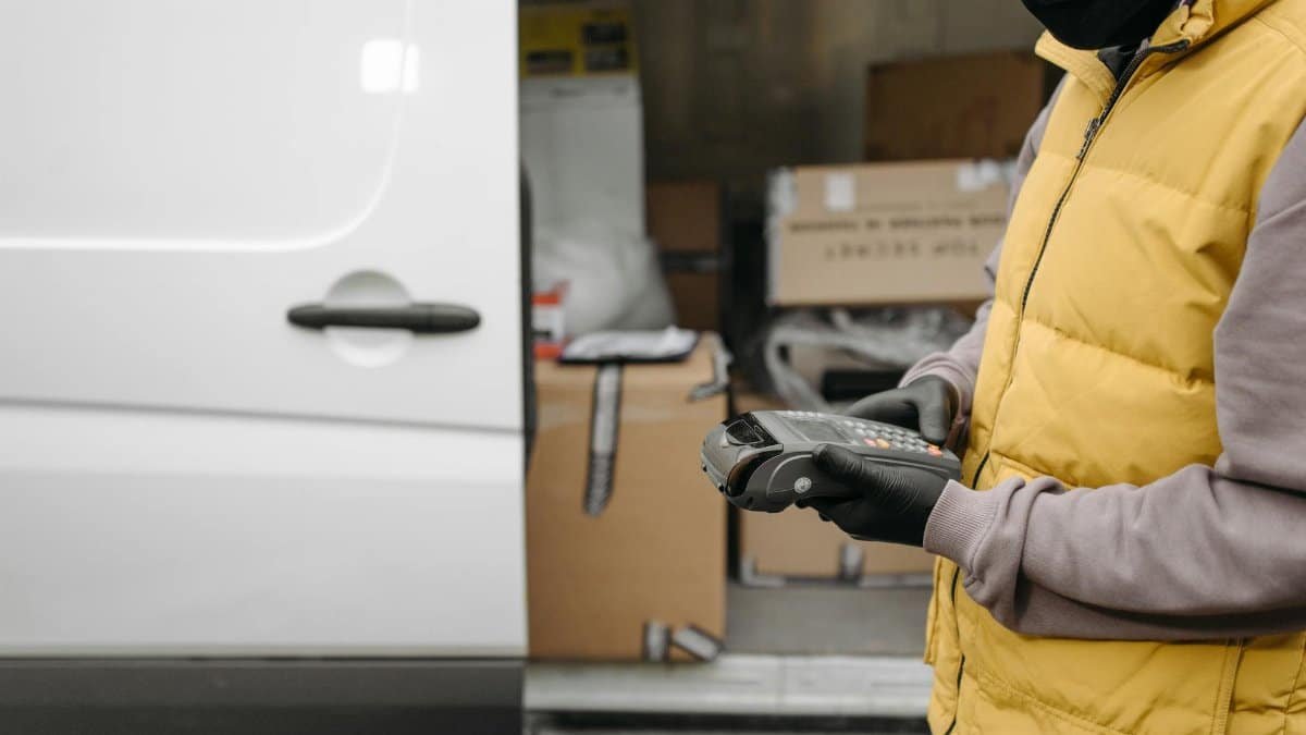 Courier using a terminal for contactless delivery payment next to a van with packages.