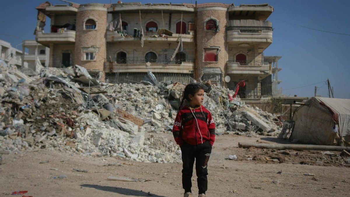 A girl stands in front of a destroyed building in Idlib, Syria, highlighting the impact of conflict.