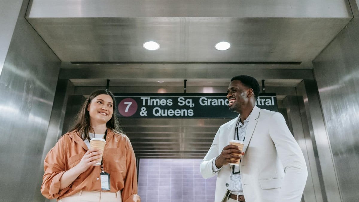 Two colleagues laugh and hold coffee near a subway sign in an underground station.