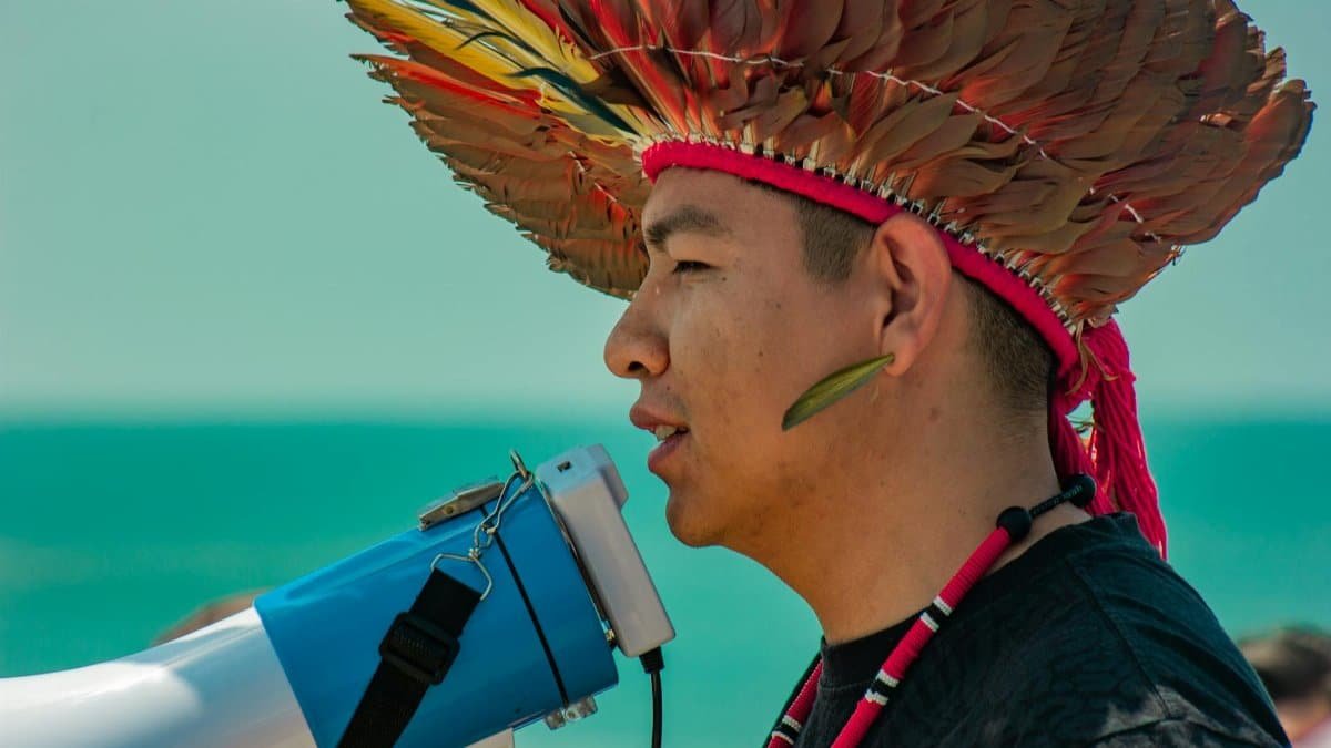 An indigenous man wearing a traditional feather headdress speaks into a megaphone outdoors in Brasil Novo, Brazil.