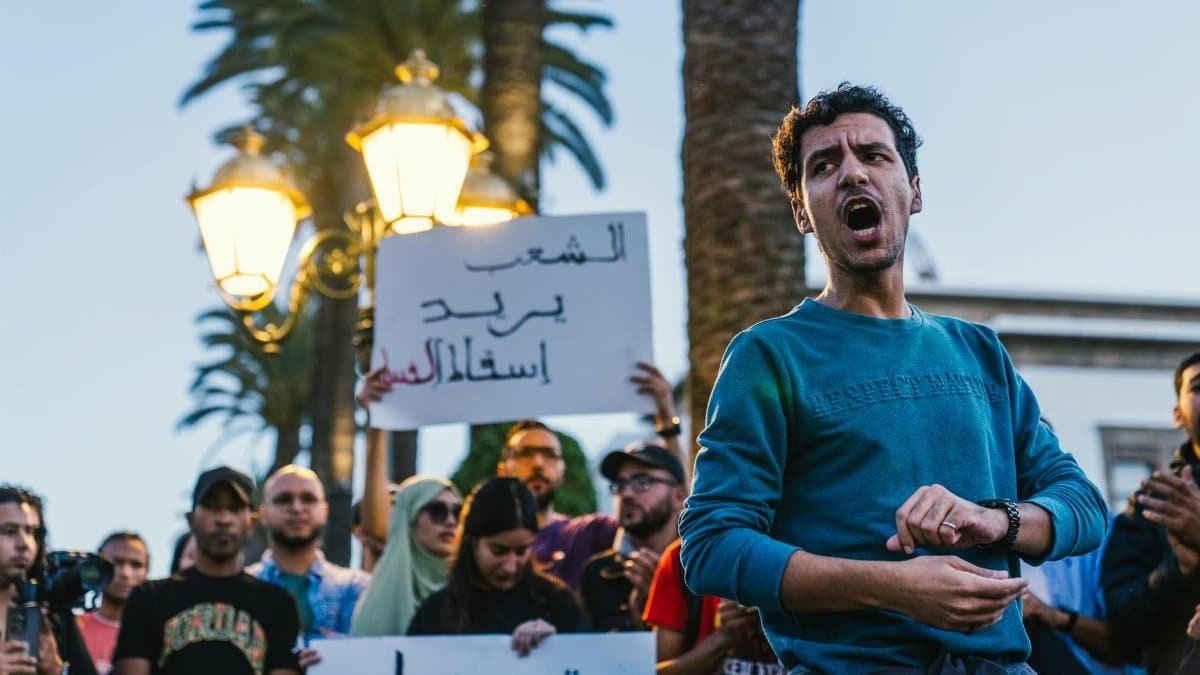 A vocal protester leads a group demonstration outdoors, holding signs with Arabic text.