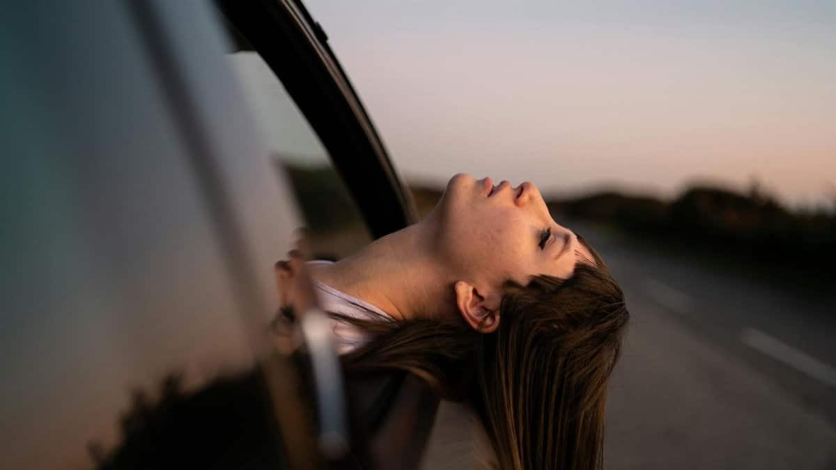 A woman leans out of a car window, enjoying a sunset drive on a quiet road.