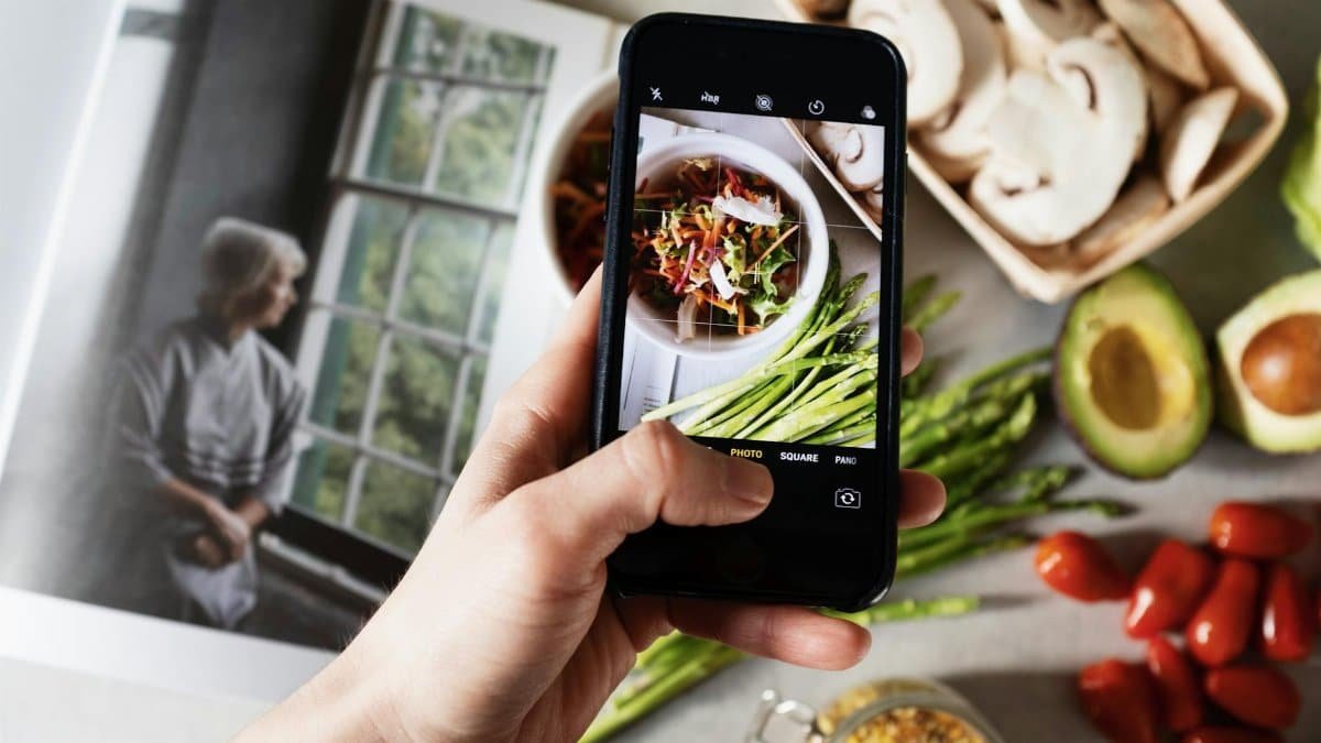 From above view of faceless blogger taking photo on smartphone of fresh tasty salad in white bowl and food placed around on table in modern kitchen