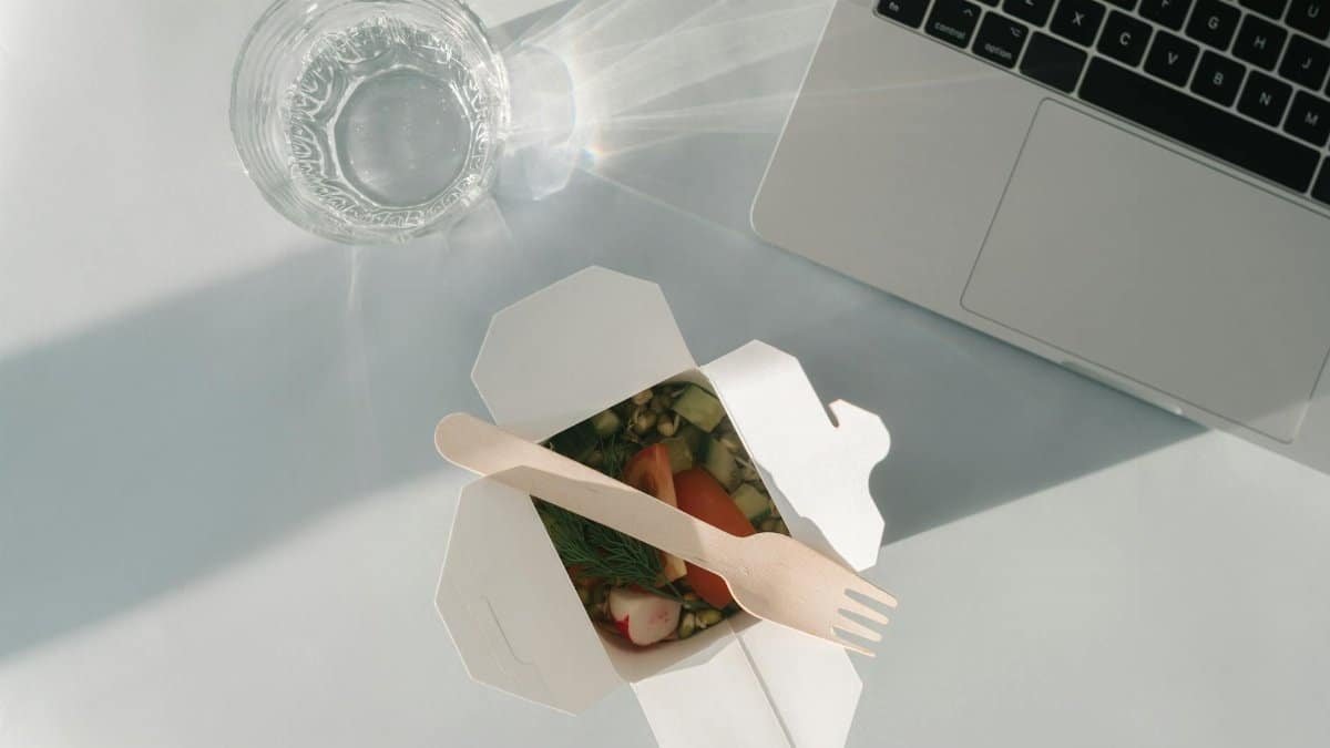 Healthy salad in a takeout box with a glass of water and a laptop on a white surface, top view.