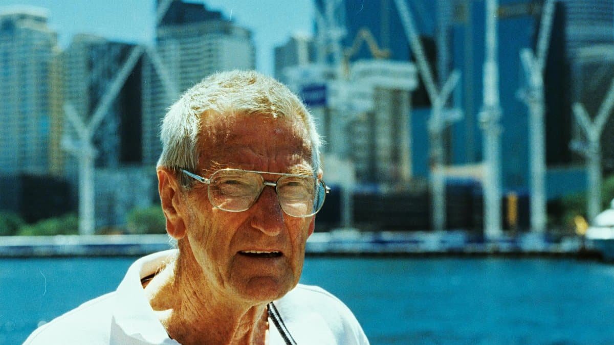 Elderly man wearing glasses at Sydney Harbour with skyscrapers in the background.