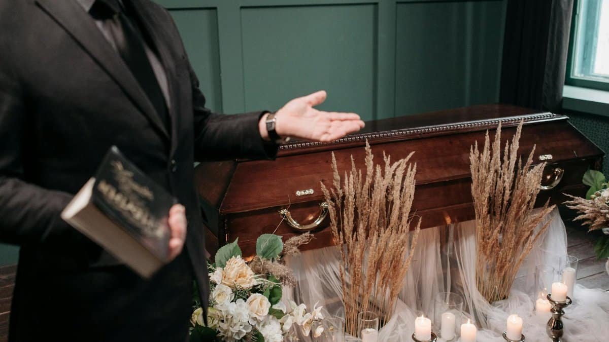 A respectful funeral setting with a closed coffin, flowers, and a man holding a book.