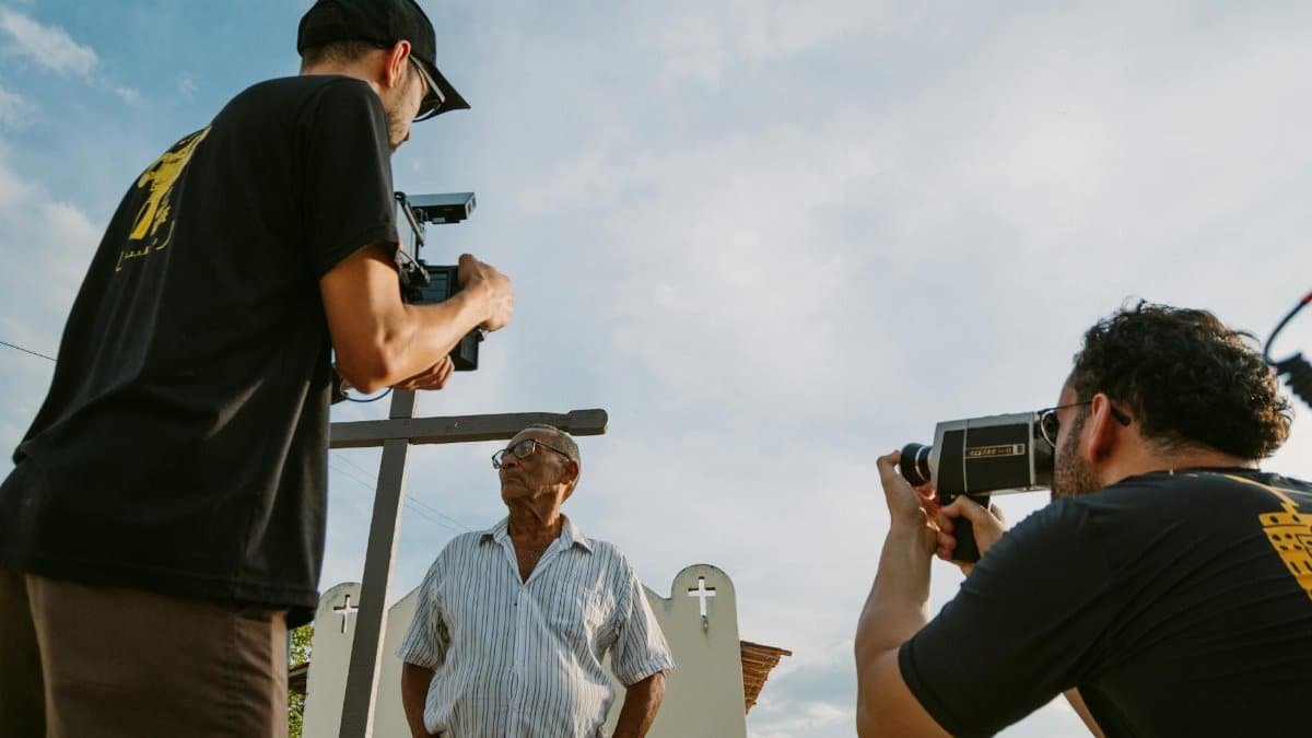 Two filmmakers document an elderly man in an outdoor setting, capturing the moment with professional cameras.