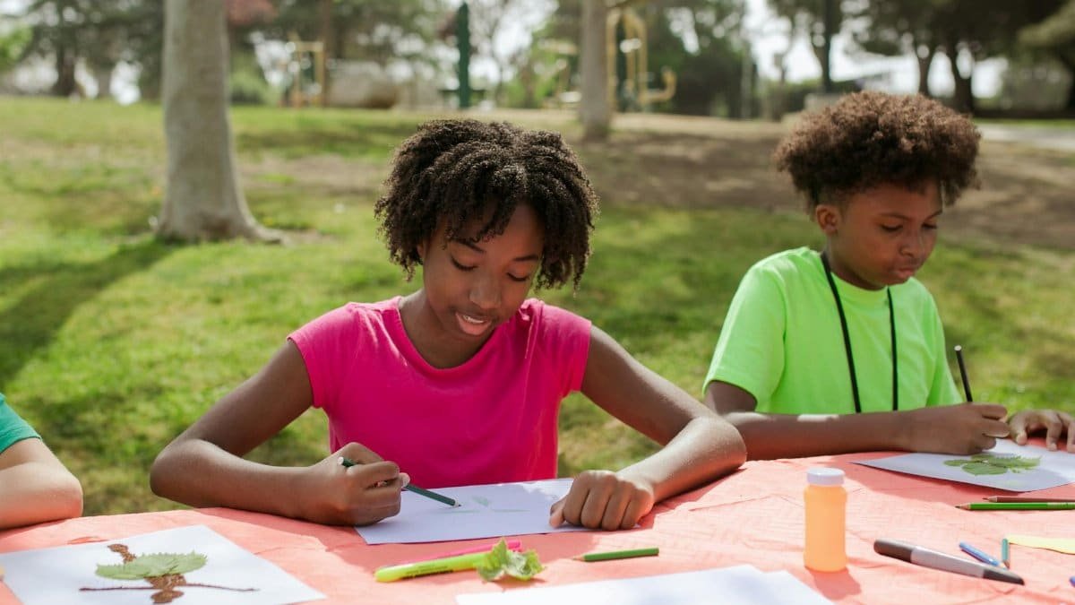Two children engage in creative drawing activities outdoors in a sunny park setting.