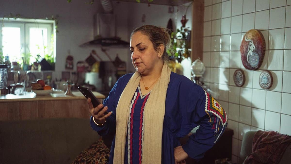 Middle-aged woman in blue dress using smartphone in a warm, cozy kitchen setting.