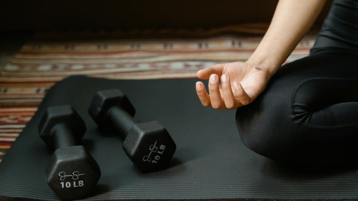 Close-up of a person's hand meditating next to dumbbells on a yoga mat, symbolizing fitness and mindfulness.