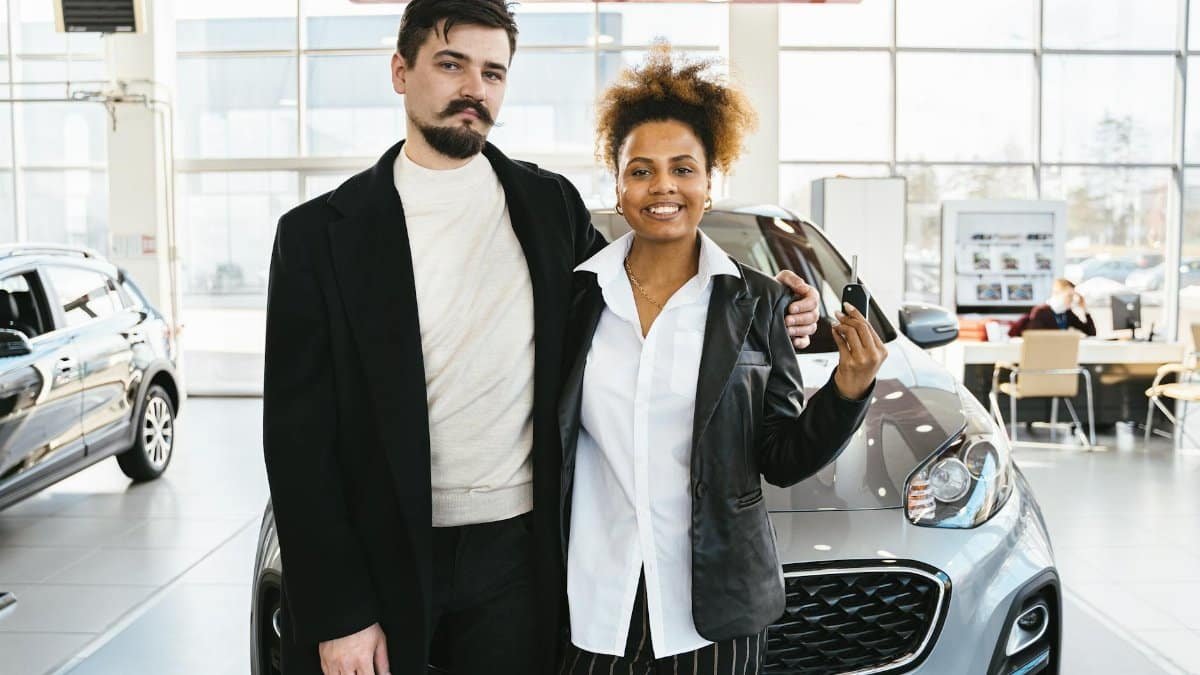 Interracial couple celebrates buying a new car at a dealership, holding keys and smiling.