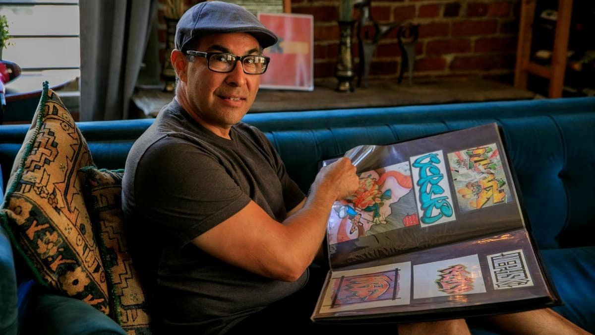 Man in hat and glasses proudly displaying his colorful graffiti art portfolio on a couch.