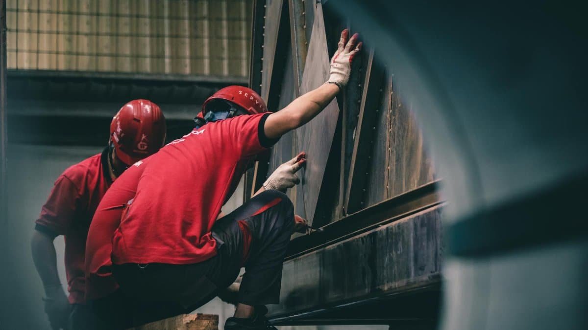 Two workers in red helmets assembling metal components inside a factory.