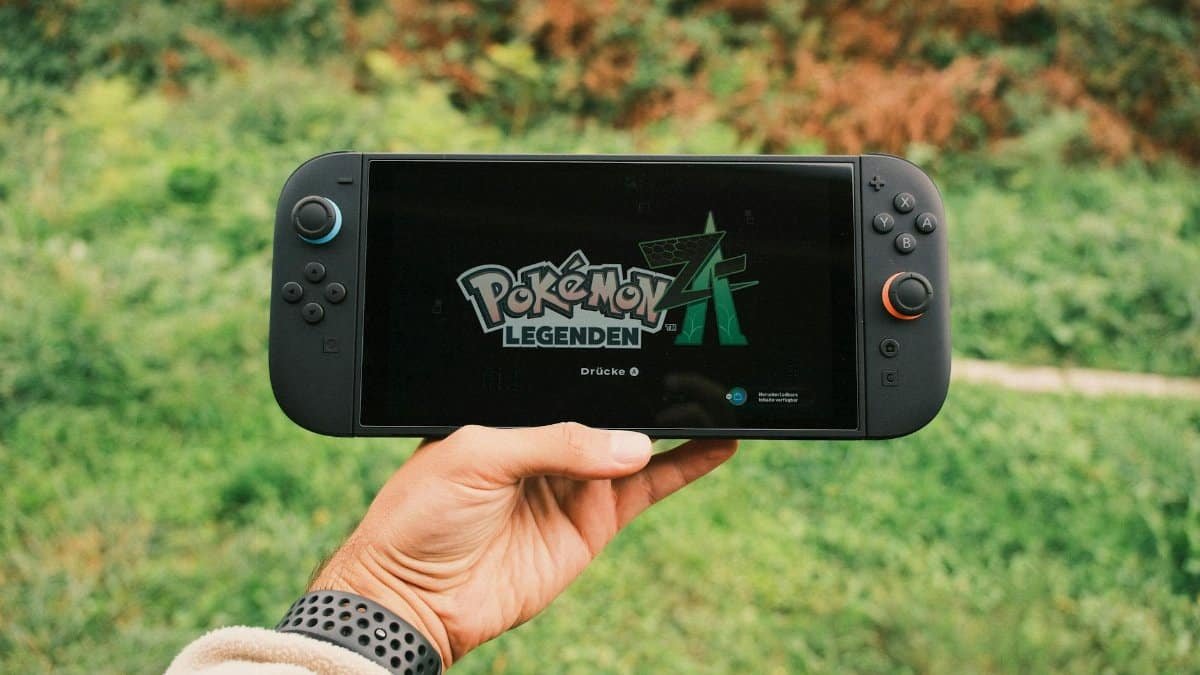A hand holding a handheld gaming console displaying the Pokémon Legends game screen outdoors.