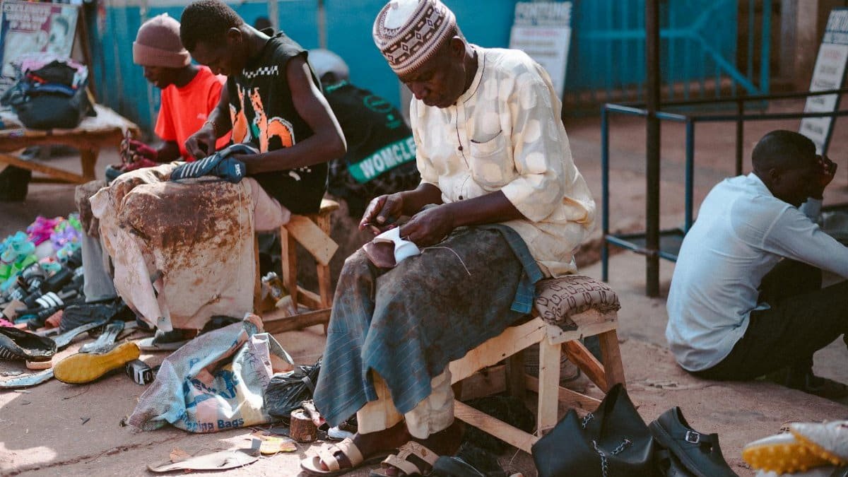 Three shoemakers crafting leather shoes in a vibrant outdoor market in Jos, Nigeria.