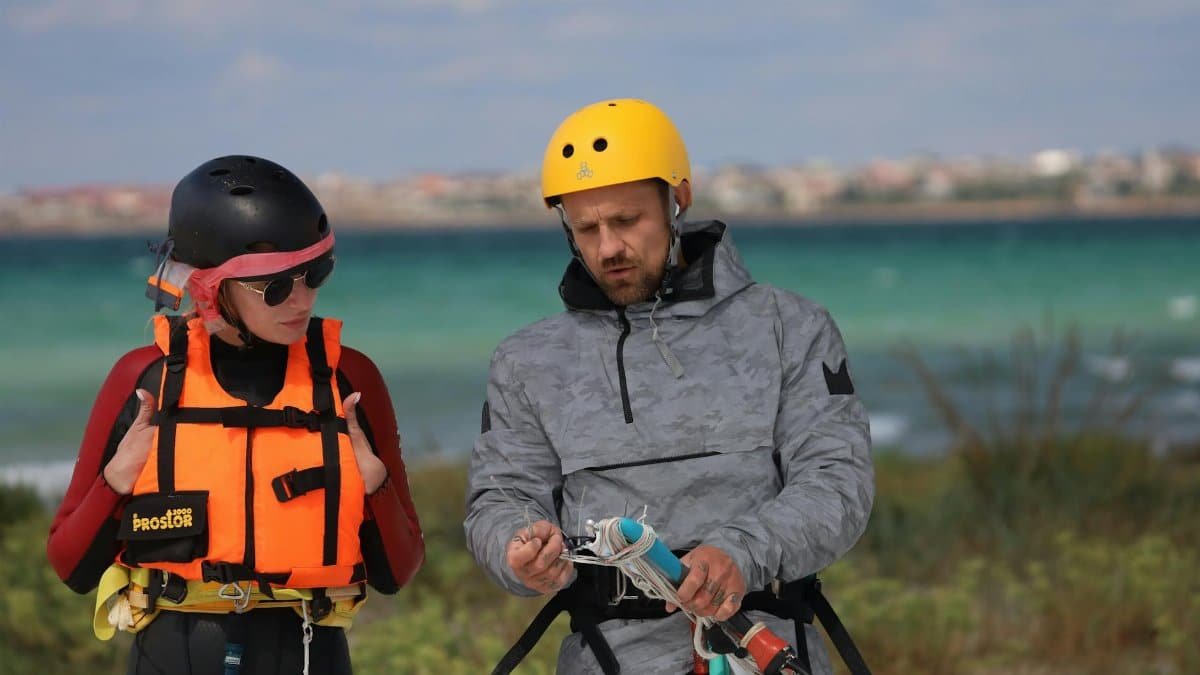 Two adults in helmets and life vests receive kitesurfing instruction on a beach.