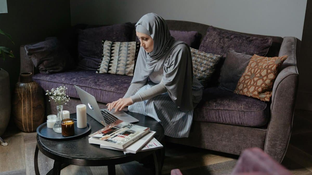 A Muslim woman in traditional attire working on her laptop in a cozy living room setting.