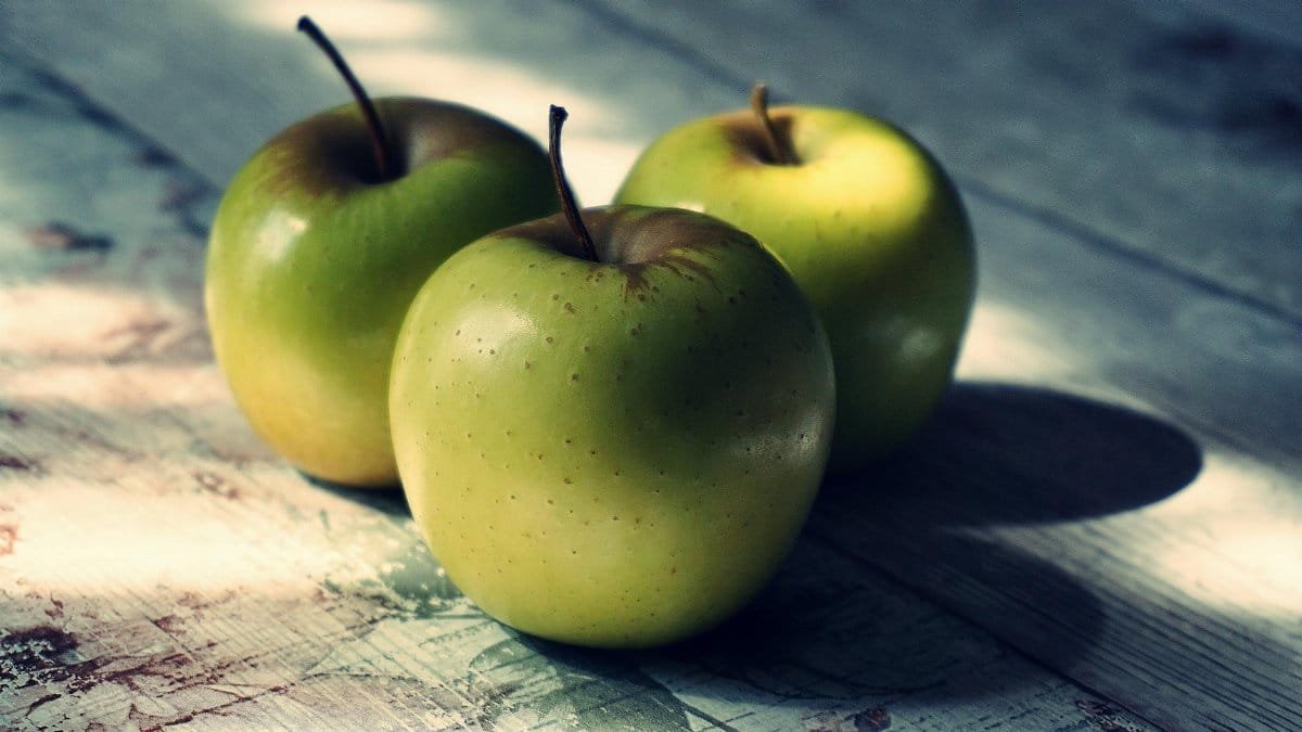 Three fresh green apples on a rustic table with natural light and shadow play.